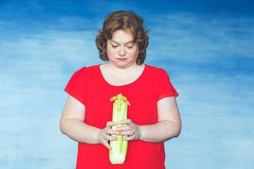 Cute smiling chubby red-haired woman looks dejectedly on celery in her hands against a blue...