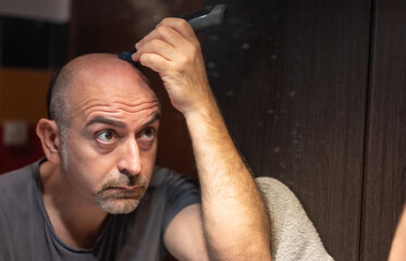 A middle-aged Caucasian man cuts his hair in the bathroom. While placing the clipper on your head check with your eyes in the mirror.