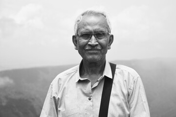 Fototapeta premium Monochrome portrait of an old man, wearing spectacles and blue shirt, standing alone in nature a blurry mountain background. The elder person with side bag on his shoulder, looking towards the camera.