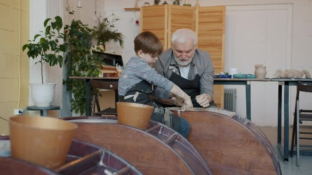 Grandfather is teaching little boy in pottery class using throwing-wheel working with clay in modern arts studio. Family hobby and occupation concept.