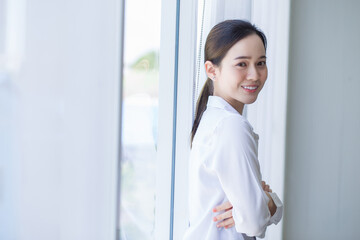 Asian beautiful woman has black long hair in white shirt. She is smiling and standing near window with white curtain.
