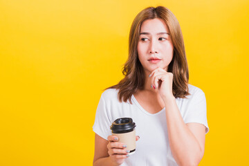 Portrait Asian Thai beautiful happy young woman standing smiling holding take away coffee paper cup she holding chin and thinking, studio shot isolated on yellow background, with copy space