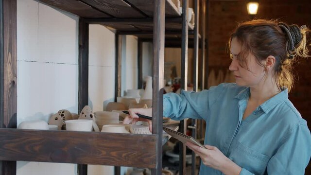Young woman is checking hand-made pots and taking notes in pottery studio working at family business. Accountancy, inventory and people concept.