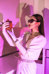 a stylish girl in black sunglasses and gloves and a white dress and a cake in her hands on a pink background