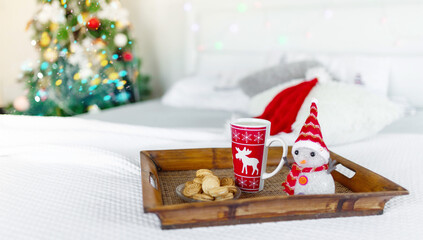 Christmas background. Red and white mug and plate with cookies, on a wooden tray, on white covered bed, near the new year tree. The concept of a cozy home evening