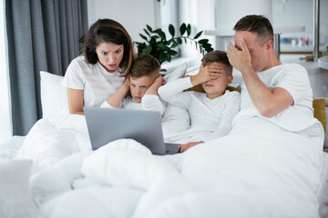 Fototapeta premium Beautiful parents with kids enjoying at home. Young family watching movie on lap top