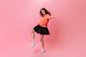 Curly girl in stylish outfit dancing on pink background