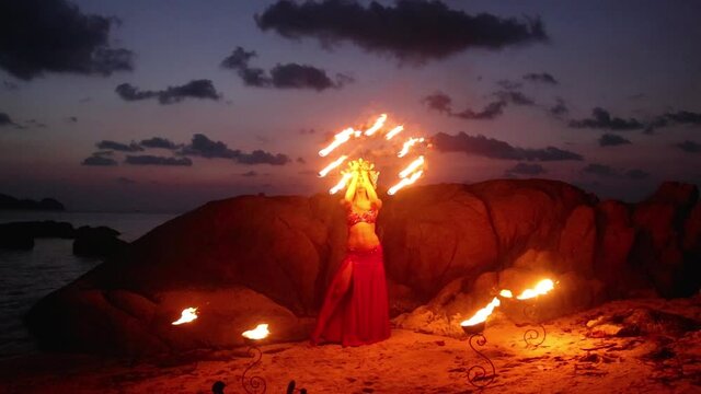 Erotic and sensual belly dancer in red costume, performing with fire props on the beach during golden hour. Slow motion.