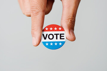 closeup of a young caucasian man with a vote badge for the United States election in his hand, on an off-white background with some blank space
