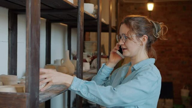 Cheerful girl is talking on mobile phone holding hand-made ceramics in pottery studio discussing family business. Communication and craft concept.
