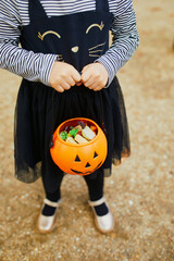 Adorable toddler girl in black cat dress with tutu skirt trick-or-treating with orange pumpkin bucket