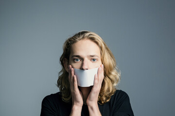 Handsome young man with long blonde hair covered his mouth with a white sheet on a gray background.