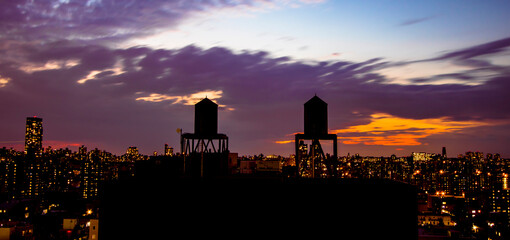 Die Silhouette von zwei Wassertanks über den Dächern Harlems am späten Abend. Der Himmel ist wolkig und in sattem blau und orange gefärbt. Im Hintergrund die Skyline von New York City