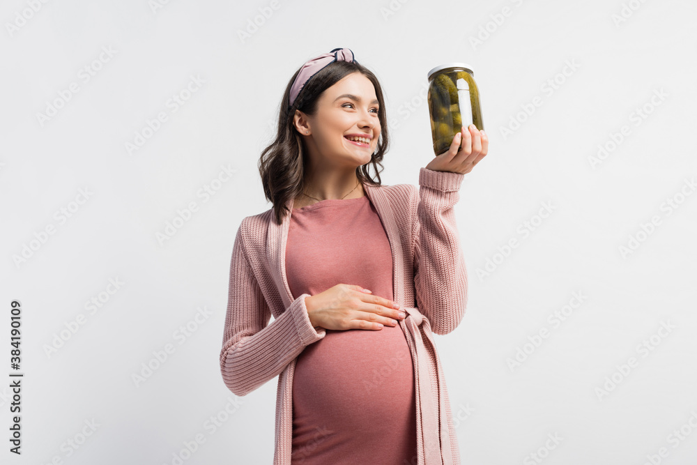 Wall mural joyful pregnant woman in headband looking at jar with pickled cucumbers isolated on white