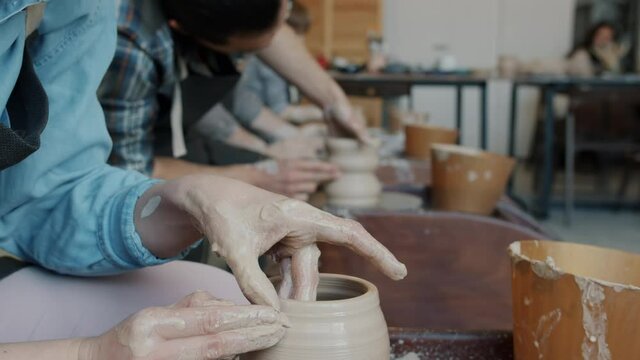 Close-up tilt-up of young woman making pot on throwing-wheel in pottery class with group of people in baackground. Hobby and leisure activity concept.