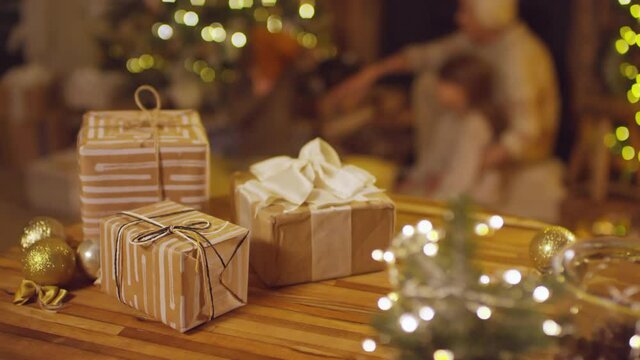 Closeup of Christmas presents wrapped in striped craft paper with bows of ribbons and golden decorations on table with grandmother with kids play in blurred background