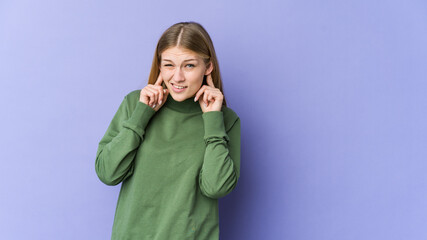 Young blonde woman isolated on purple background covering ears with fingers, stressed and desperate by a loudly ambient.