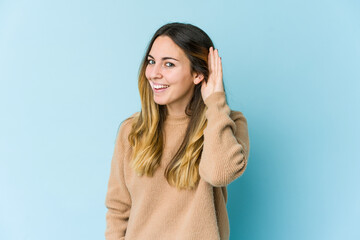 Young caucasian woman isolated on blue background trying to listening a gossip.