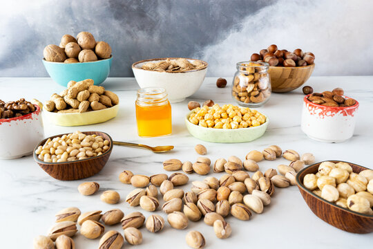 Various nuts (hazelnuts, almonds, walnuts, cashews, pine nuts) peeled and in shell in wooden and ceramic bowls are on a white table. In the center there is a small jar of honey and scatterer pistachio