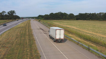Aerial shot of truck with cargo trailer driving on road and transporting goods. Flying over delivery lorry moving along highway passing in countryside with scenic nature environment around. Back view