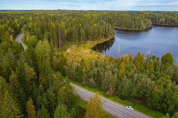 White car on the highway road. Blue lake and autumn green, yellow forest around. Aerial drone view. Karelia, 