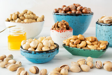 Walnuts, cashews, almonds and hazelnuts in beautiful bowls (blue, white, green) stand a white background. A honey stay in a small jar with gold spoon. 
