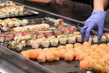 A selection of sushi in the buffet. Somebody with a violet rubber glove is organizing them.
