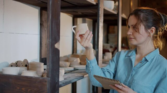 Portrait of female potter checking ceramics and writing information in notebook in workshop busy with inventory. People and occupation concept.