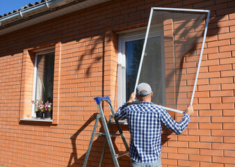 A man on a step ladder is installing a mosquito net, fly screen or frame on plastic window  to...