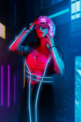 Cyberpunk pink haired woman in neon glasses and transparent raincoat with luminous stripes on night city background with neon lights. © syberianmoon