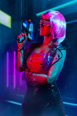 Cyberpunk pink haired woman in neon glasses and transparent raincoat holding a gun on night city background with neon lights.