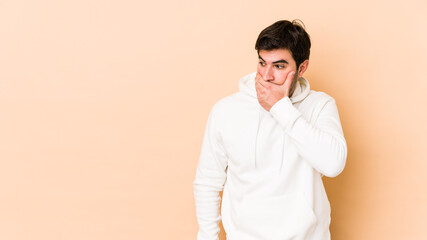 Young man isolated on beige background thoughtful looking to a copy space covering mouth with hand.