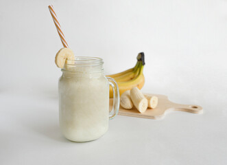 Fresh banana smoothie in a glass jar with a straw, near bananas, on a light background. Healthy food. selective focus.