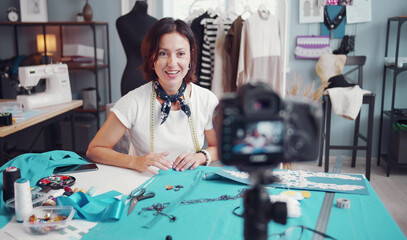 Female seamstress blogger sitting at table in workshop shooting video tutorial on how to sew clothes