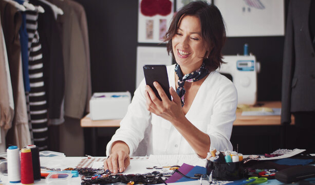 Fashion designer with beaming smile sitting at table in tailor workshop and browsing smartphone