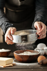 Chef hands pouring flour powder on raw dough using sieve on a black background, Cooking process.