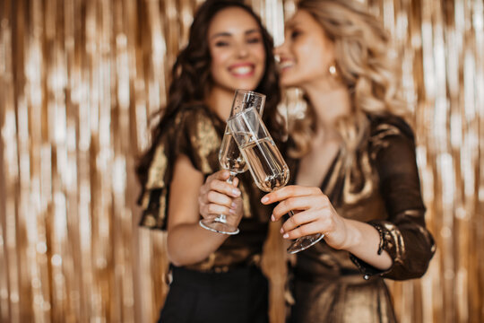 Girls in bronze-colored dresses clink glasses with champagne on gold background