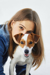 A portrait of a young confident caucasian woman in a denim jacket with her pet, jack russell terrier. She is hiding behind the happy dog, kissing him and looking into the camera. White background.