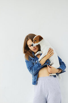 Young caucasian woman in a denim jacket is calmly holding her pet, jack russell terrier. She has her eyes closed. The dog is looking in front of him. White background.