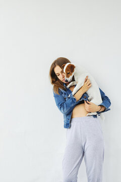 Young caucasian woman in a denim jacket is holding her pet, jack russell terrier, as if protecting the puppy. She and the dog are focused, looking down on something on the ground. White background.