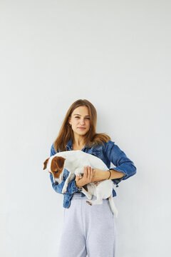 Young caucasian woman in a denim jacket is holding her pet, jack russell terrier, on white background. She is looking into the camera, the dog is focused and looks down, on the ground. 
