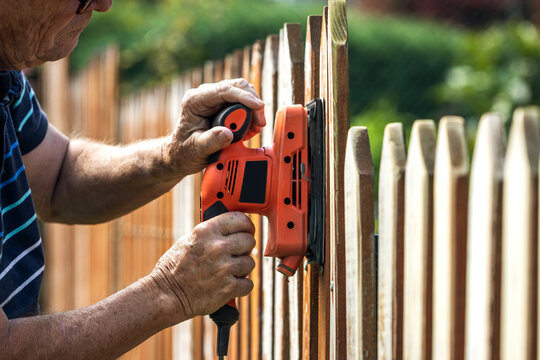 Sanding wood fence by sander. Senior man repairing picket fence in garden