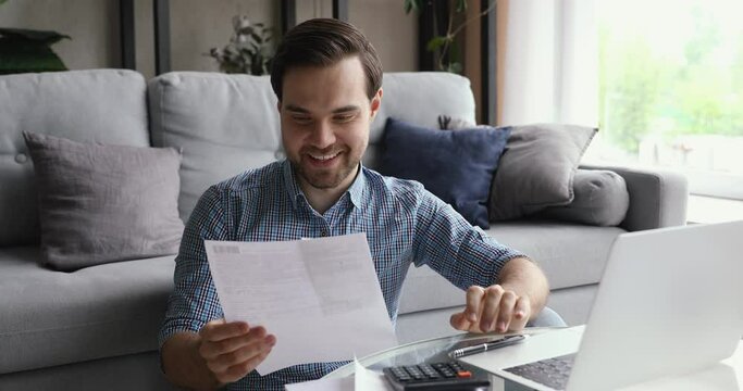 Smiling young man holding paper bills invoices in hands, managing income and outcome in computer application, making household payment online, feeling happy of enough money, positive banking balance