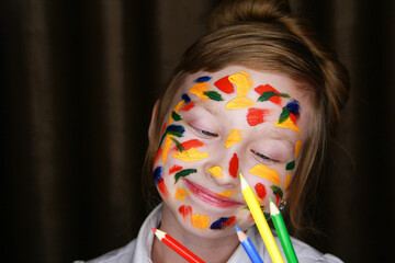 A girl stained with paint holds colored pencils.