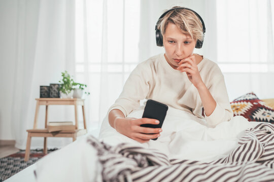 Young female sitting on cozy bed dressed pajamas browsing the internet and listening to music using wireless headphones. Music playing modern technology and free time spending concept