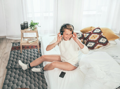Smiling young female sitting on cozy bed dressed pajamas listening to music using wireless headphones. Music playing modern technology and free time spending concept