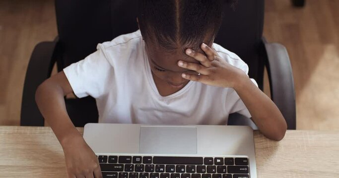 Close up little African American ethnic girl sitting at table typing on laptop keyboard feeling angry irritation problem. Mixed race female child holding her head from stress hard homework, top view