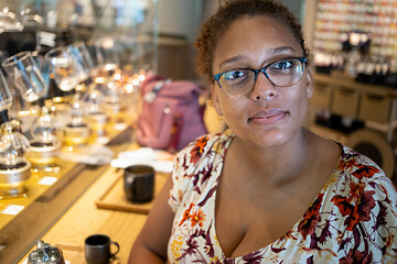 portrait of an African american black woman with glasses is sitting down in coffee cafe happily. she is wearing a colorful dress and waiting on her drink.