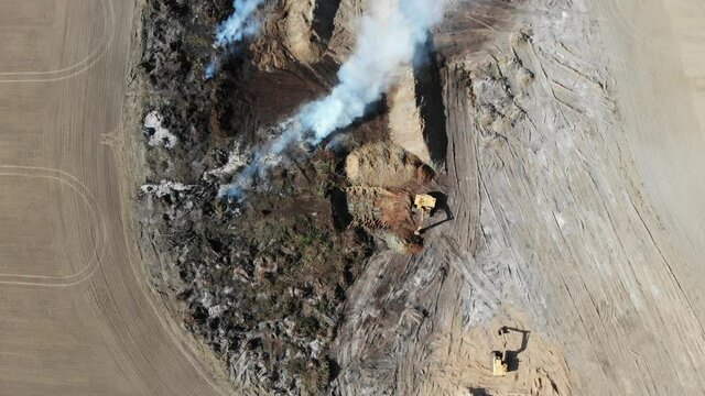 Excavators clear the field from the forest to increase farmland. Burning trees in a pit after increasing the plowed field. Inefficient farming. Environmental problem. Aerial view 4k.