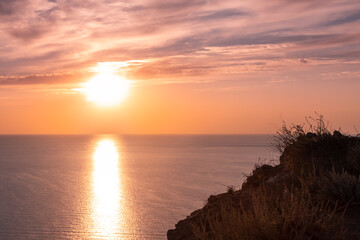 A large crimson Sun sets over the horizon against a cloudy sunset pink sky. Twilight sun shines on the top of the cliff. The edge of the cliff turns into a abyss. Black evening sea below the cliffs.